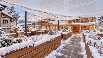 Un’area salotto esterna innevata con panche e tavoli di legno, un bar e vista sulle montagne innevate sullo sfondo.