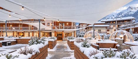 A snow-covered courtyard with wooden planters, a central pathway, and buildings with balconies and windows.