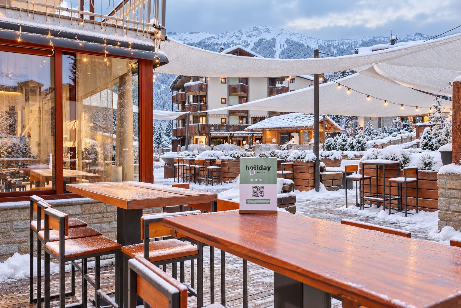 An outdoor seating area with wooden tables and chairs, a sign indicating a holiday promotion, and a view of snow-covered buildings in the background.