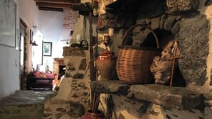 A rustic stone interior with a stone fireplace, shelves with pottery, and a wooden staircase.