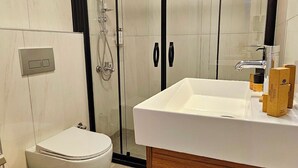 Uma casa de banho moderna com chuveiro em cabine de vidro, lavatório branco com base de madeira, sanita e um lixo.