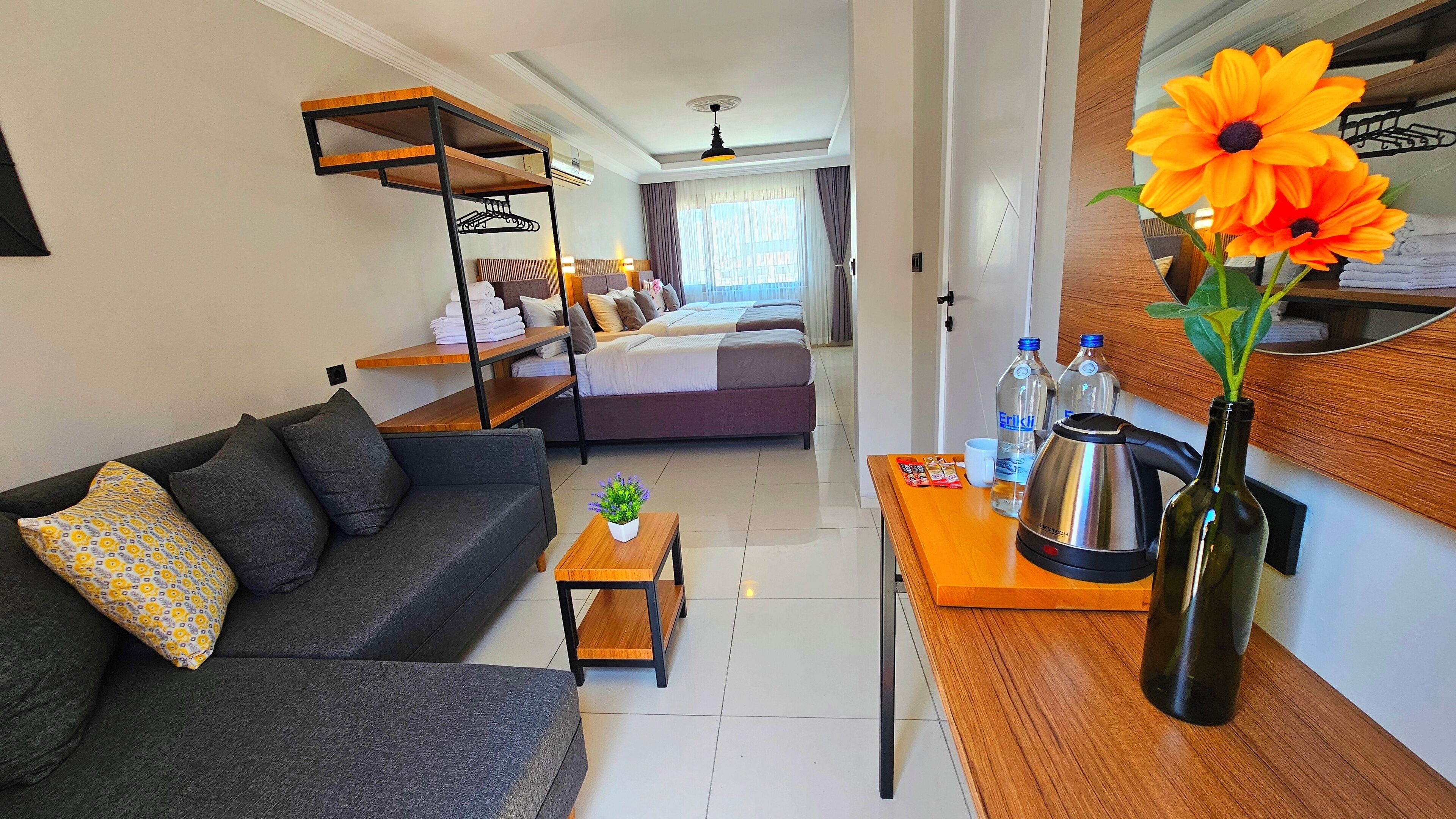 A modern hotel room with a sofa, a bed with a headboard, a small table with a plant, and a coffee maker.