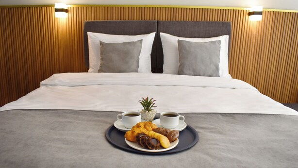Cama bem arrumada com manta cinza, duas almofadas e bandeja com doces e xícaras de café.