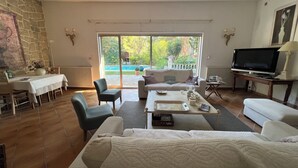 A living room with a stone wall, a dining area, a sofa, a TV, and a view of a pool and greenery outside.