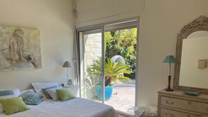 A bedroom with a bed, pillows, a nightstand, a dresser, a mirror, and a large window with a view of greenery outside.