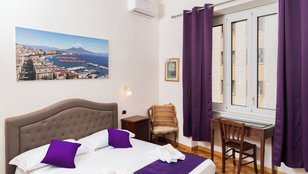 A hotel room with a bed, a desk, and a chair. There is a painting on the wall and a window with purple curtains. - Suite Cervantes (Naples)