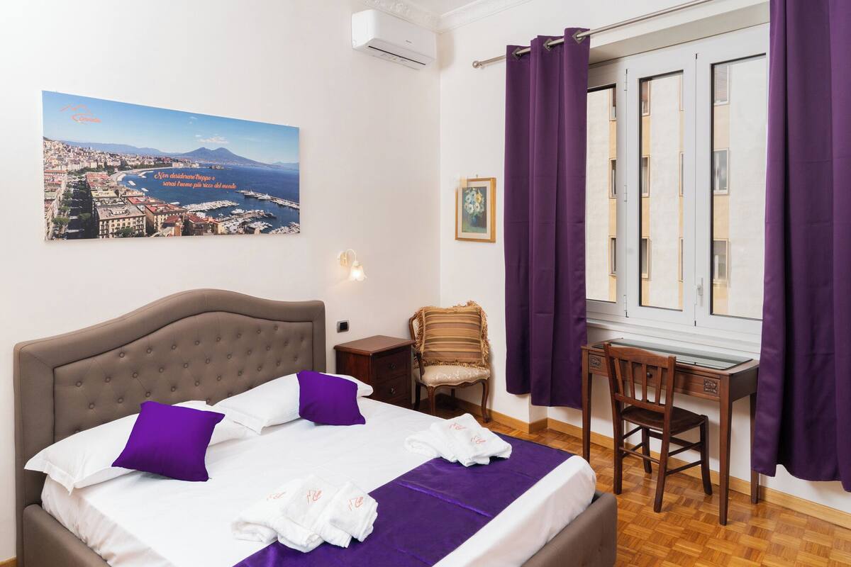 A hotel room with a bed, a desk, and a chair. There is a painting on the wall and a window with purple curtains.
