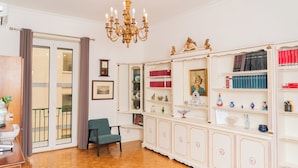 A room with a wooden floor, a green chair, a white cabinet with red and blue books, a chandelier, and a window with curtains. - Suite Cervantes (Naples)