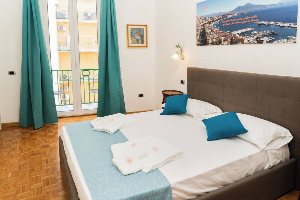 A hotel room with a bed, a view of a coastal city, and a balcony with green curtains.