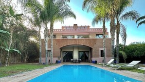 A swimming pool with lounge chairs, surrounded by palm trees and a modern building with large windows. - Villa in the heart of Marrakech (Bab Ighl Marrakesh,)