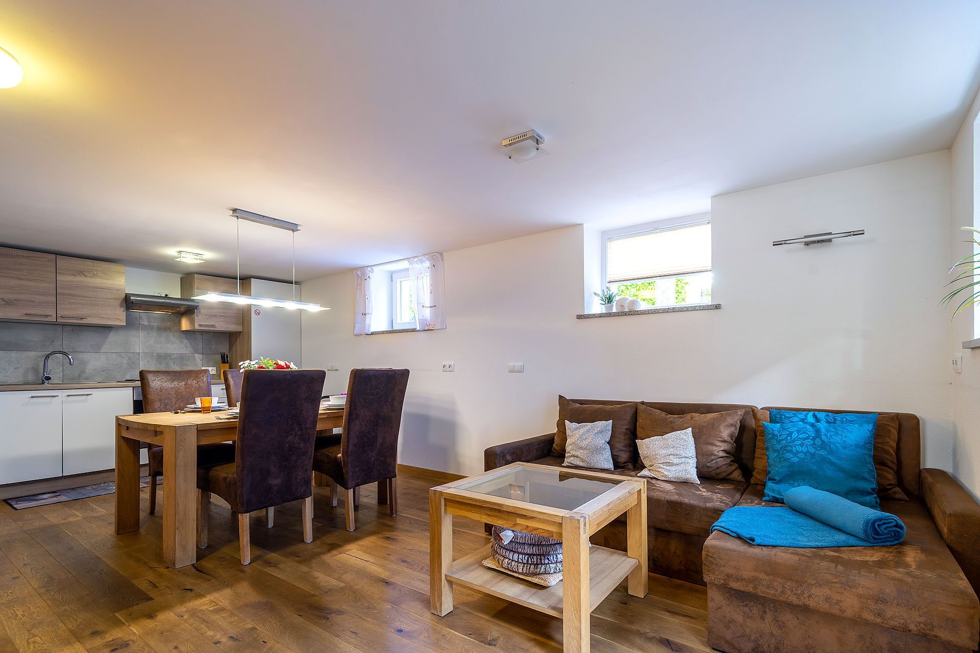 A modern living room with a wooden dining table, dark chairs, a glass coffee table, and a brown sofa with cushions.