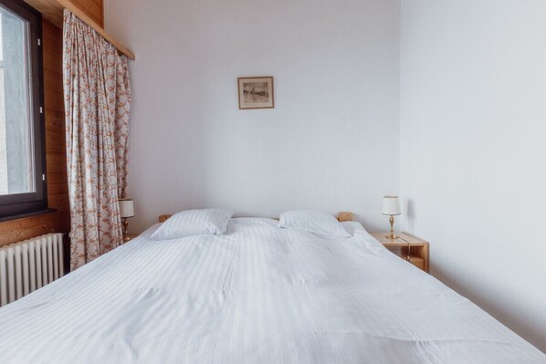 Una cama bien hecha con sábanas blancas, un cabecero de madera, una mesita de noche con una lámpara, y una ventana con cortinas.