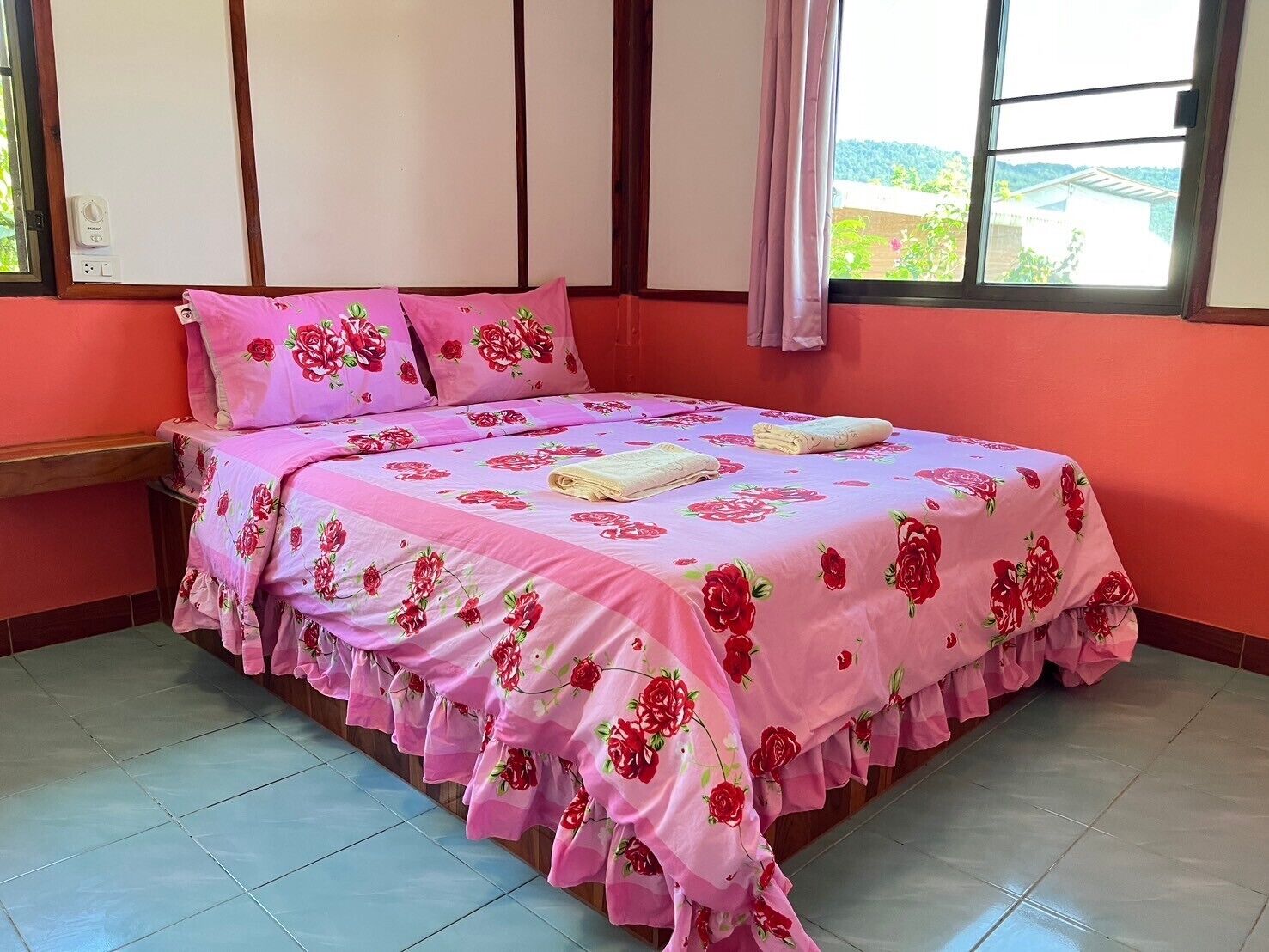A bedroom with a bed covered in pink floral bedding, a window with a view of greenery, and a wooden headboard.