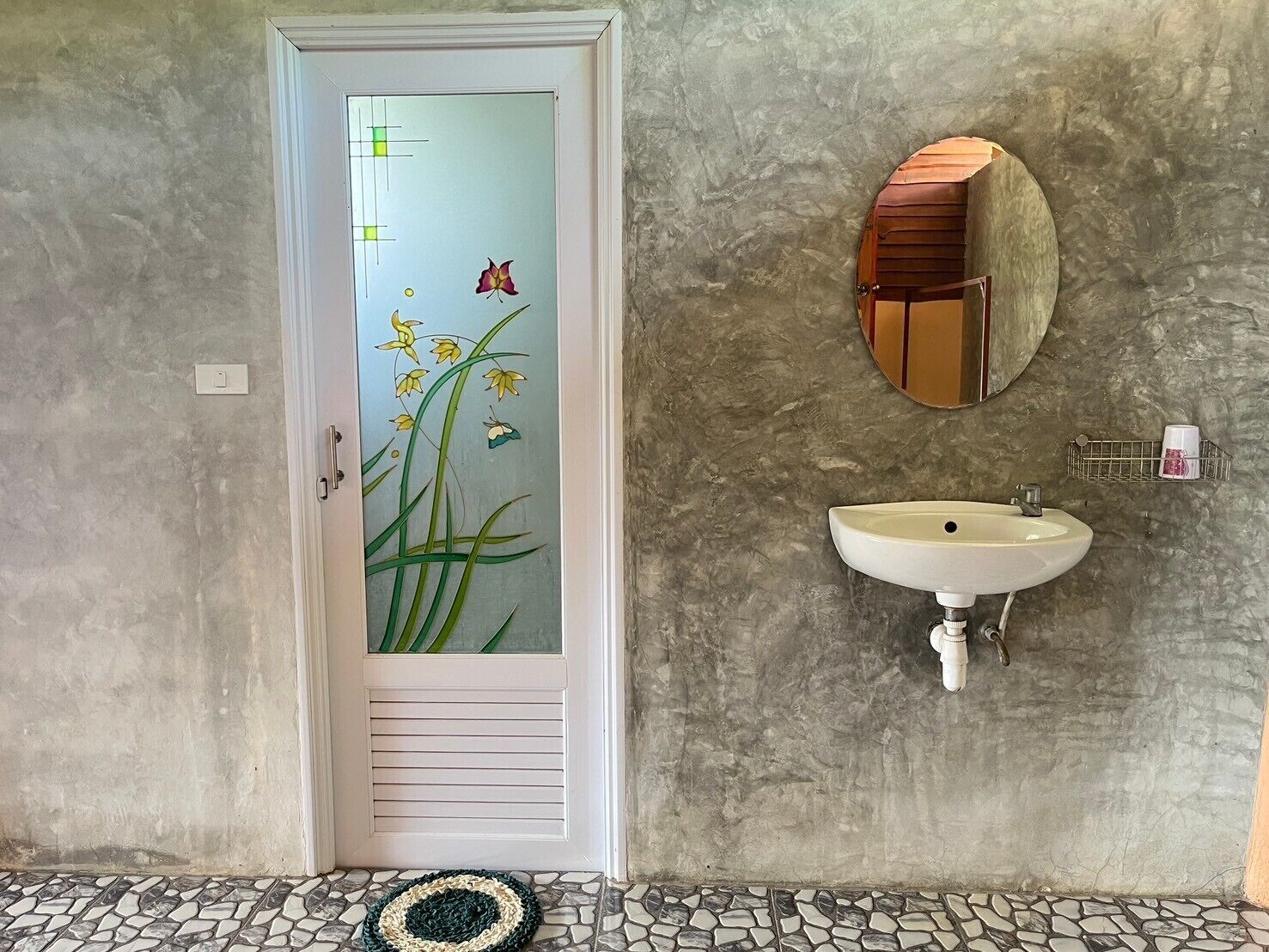 Une salle de bain avec une porte blanche, un miroir rond, un lavabo blanc et un sol carrelé.