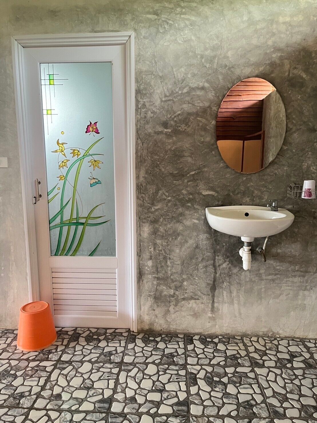 A bathroom with a stone tile floor, a white sink, a mirror, and a door with floral glass.