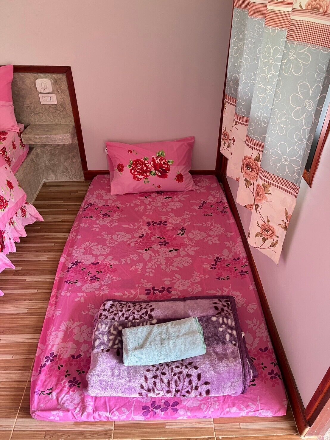 A small, single-bed room with floral bedding, a wooden floor, and a window with floral curtains.
