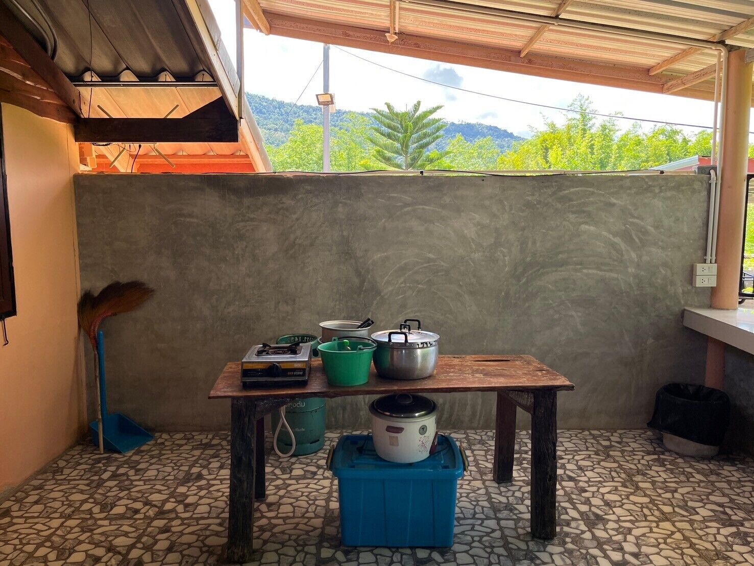 Un pequeño espacio de cocina con una mesa que sostiene ollas y una estufa a gas, un recipiente azul y un cubo verde.