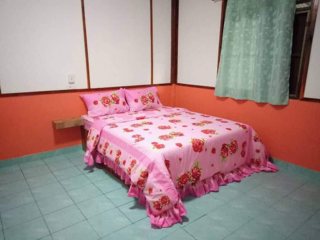 Un dormitorio con una cama cubierta por una colcha floral rosa, una mesita de noche de madera y una cortina verde.