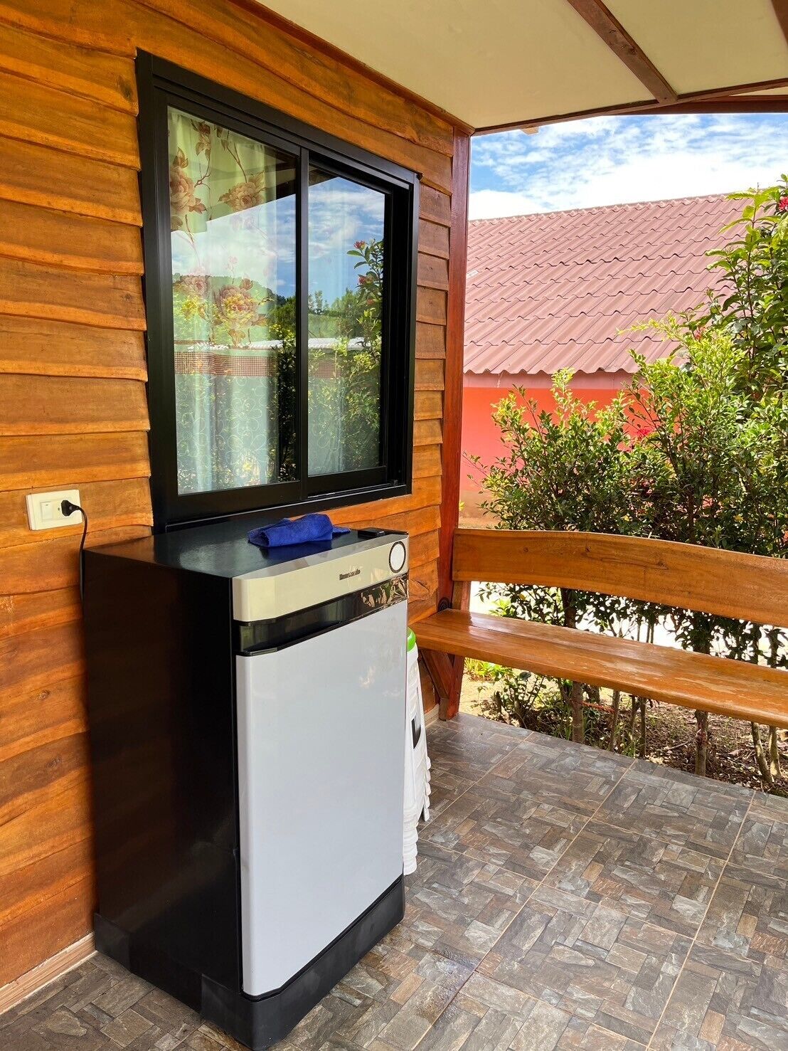 A compact kitchenette with a built-in refrigerator, a window with a floral curtain, and a view of a red-roofed building and greenery.