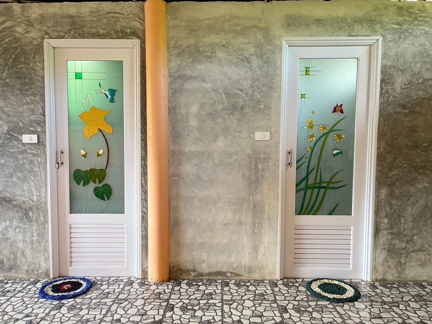 Dos puertas con paneles de vidrio esmerilado que presentan diseños florales, una pared con textura y un suelo de baldosas con dos felpudos.