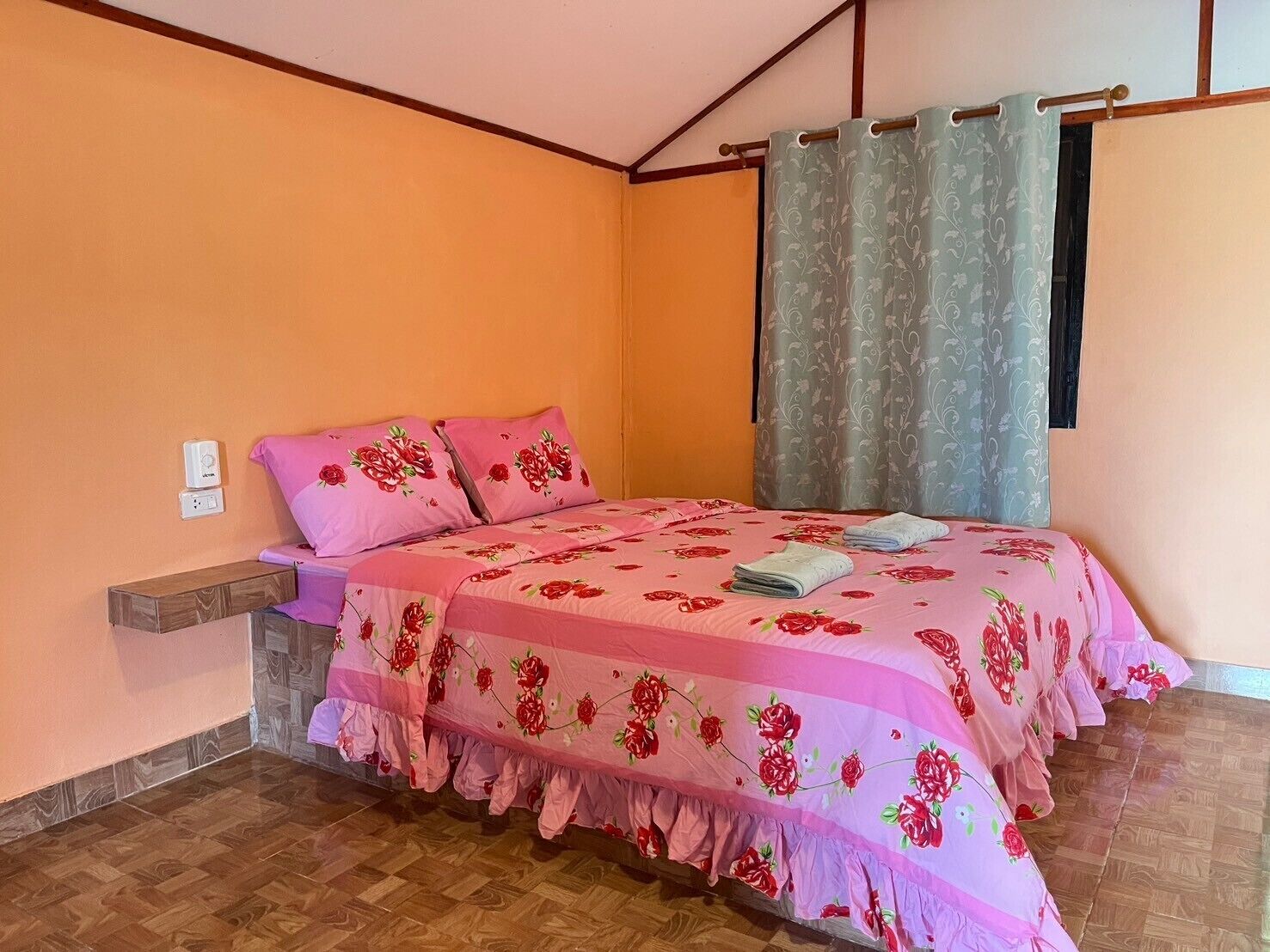 Un dormitorio con cama, mesita de noche de madera, enchufe y ventana con cortinas.