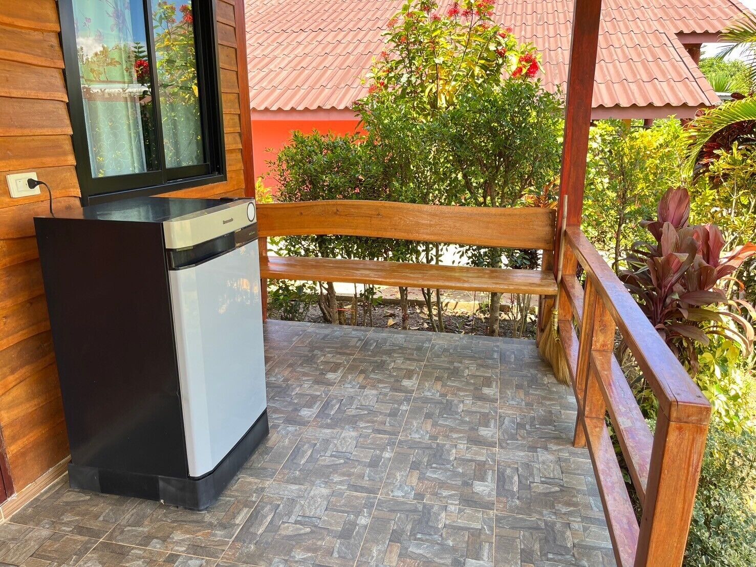 A compact kitchenette with a refrigerator and a small countertop, a wooden bench, and a tiled patio area with surrounding greenery.