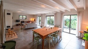 Ein geräumiger Wohnbereich mit einem Holzesstisch, grünen Stühlen, einer Couch und einem Fernseher. Große Fenster sorgen für natürliches Licht und einen Blick ins Grüne.