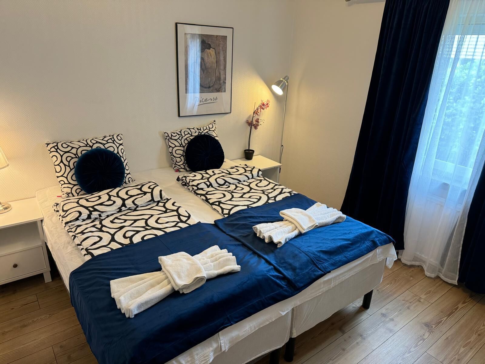 A double bed with patterned bedding, a bedside table with a lamp, a framed artwork on the wall, and a window with sheer curtains.