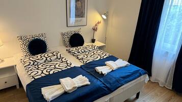 A double bed with patterned bedding, a bedside table with a lamp, a framed artwork on the wall, and a window with sheer curtains.