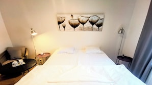 Una cama bien tendida con sábanas blancas, una mesita de noche con lámpara, una silla y una obra de arte colgada en la pared sobre la cama.