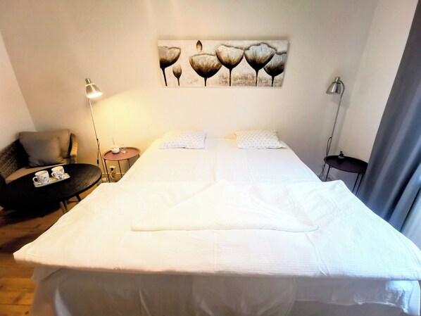 A neatly made bed with white linens, a small bedside table with a lamp, a chair, and a wall-mounted artwork above the bed. - Lamar hotel (Angelholm)