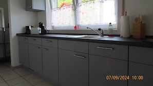 Een keuken met grijze kastjes, een roestvrijstalen spoelbak en een raam met bloemen gordijnen.