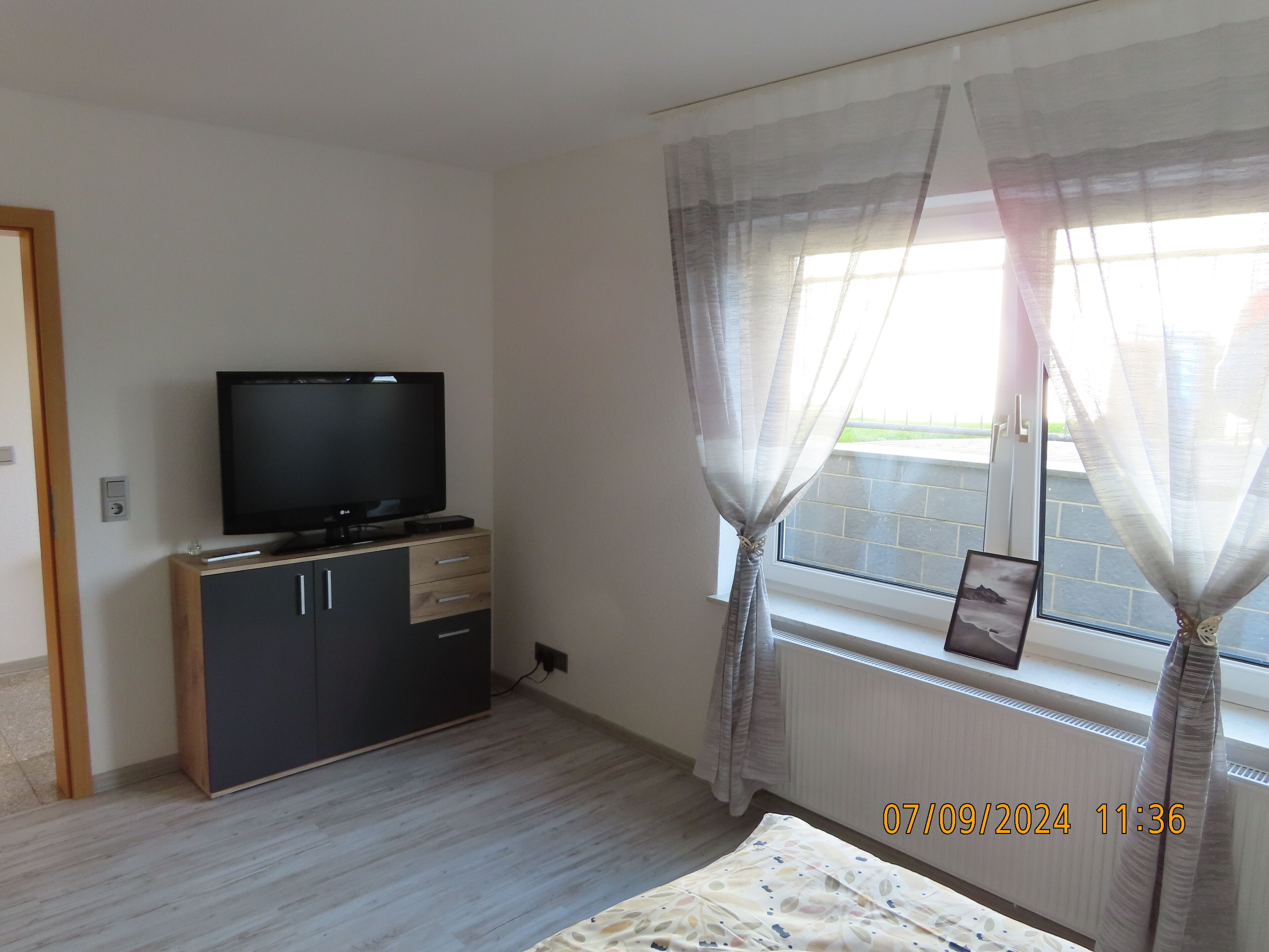 A bedroom with a TV on a wooden cabinet, a bed with a patterned bedspread, and a window with sheer curtains.