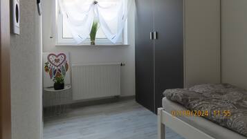 Un dormitorio con cama, armario, radiador, ventana con cortinas y una mesita con planta.