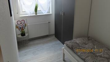 Ett sovrum med en säng, en garderob, ett element, ett fönster med tunna gardiner och ett litet bord med en växt.
