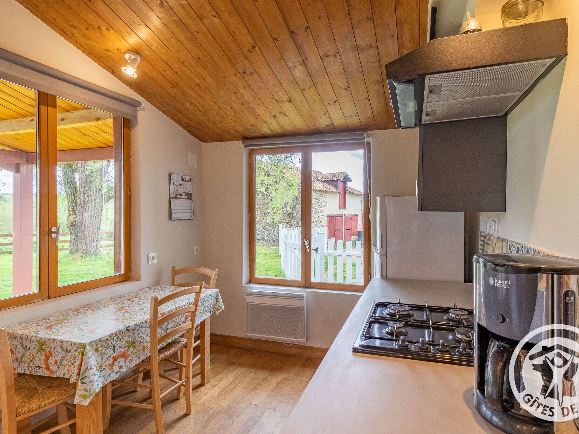 Une cuisine avec un plafond en bois, une cuisinière à gaz, une machine à café et une fenêtre donnant sur un bâtiment rouge et des arbres.