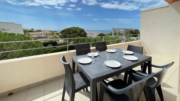 Un área de comedor al aire libre con una mesa dispuesta para cuatro personas, rodeada de sillas y vista a un paisaje urbano con edificios y un árbol.