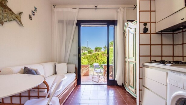 Un espacio habitable compacto con sofá, mesa de comedor, sillas, una cocina con lavadora y un ventanal corredero que da a un área exterior.