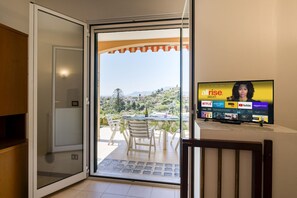 A room with a sliding glass door leading to an outdoor patio with a table and chairs, a television on a stand, and a view of a hilly landscape.
