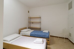 A small, clean room with two beds, a metal shelf unit, and a white chair.