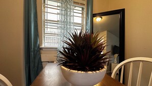 Et spisebord med en hvid skål med en plante, et vindue med persienner og et spejl på væggen.