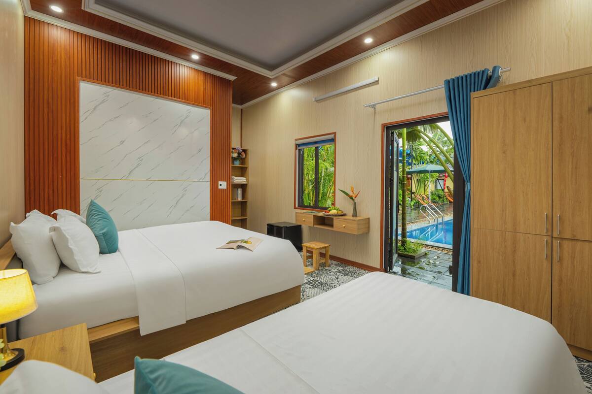 A hotel room with two beds, a wooden wardrobe, a small table with a lamp, a shelf with decorative items, and a view of an outdoor pool area.