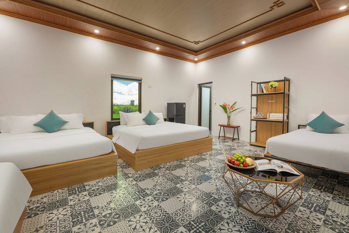 A hotel room with two beds, a small table with fruits and books, a bookshelf, and a door leading to a bathroom.
