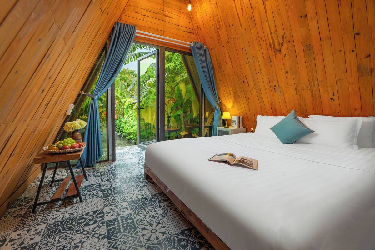 A bedroom with a large bed, a nightstand, a small table with fruit, and a view of greenery through the window.