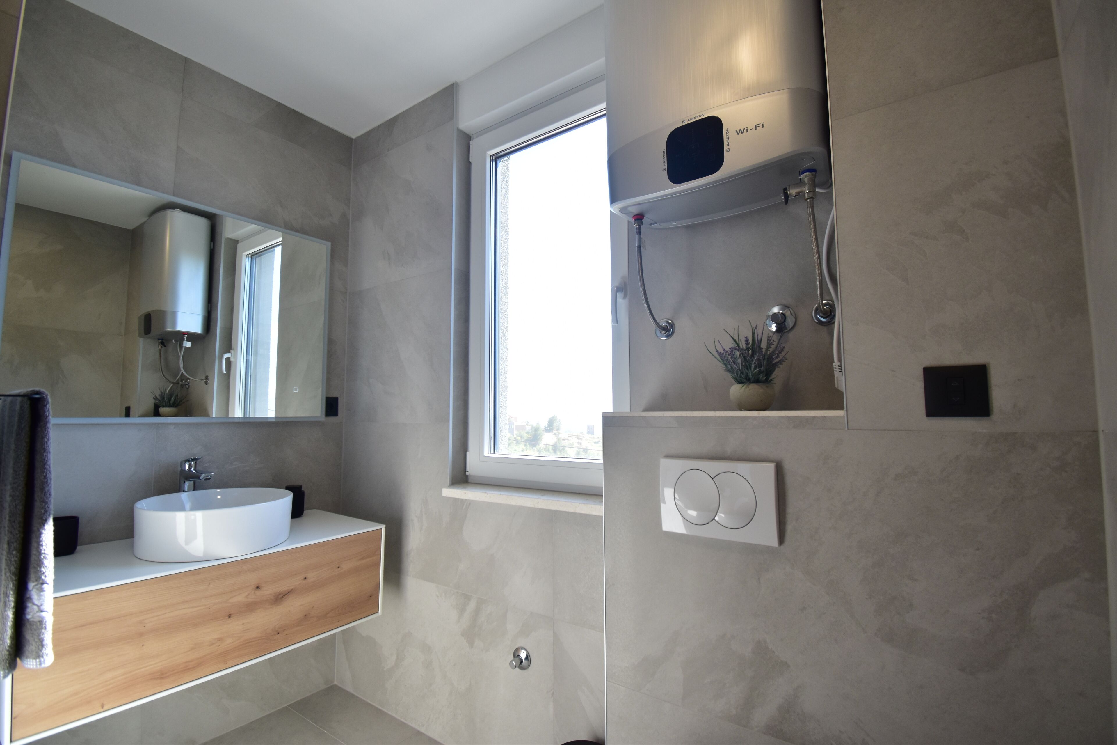 Un bagno moderno con uno specchio grande, un lavandino bianco, una doccia con soffione manuale e una finestra che offre luce naturale.