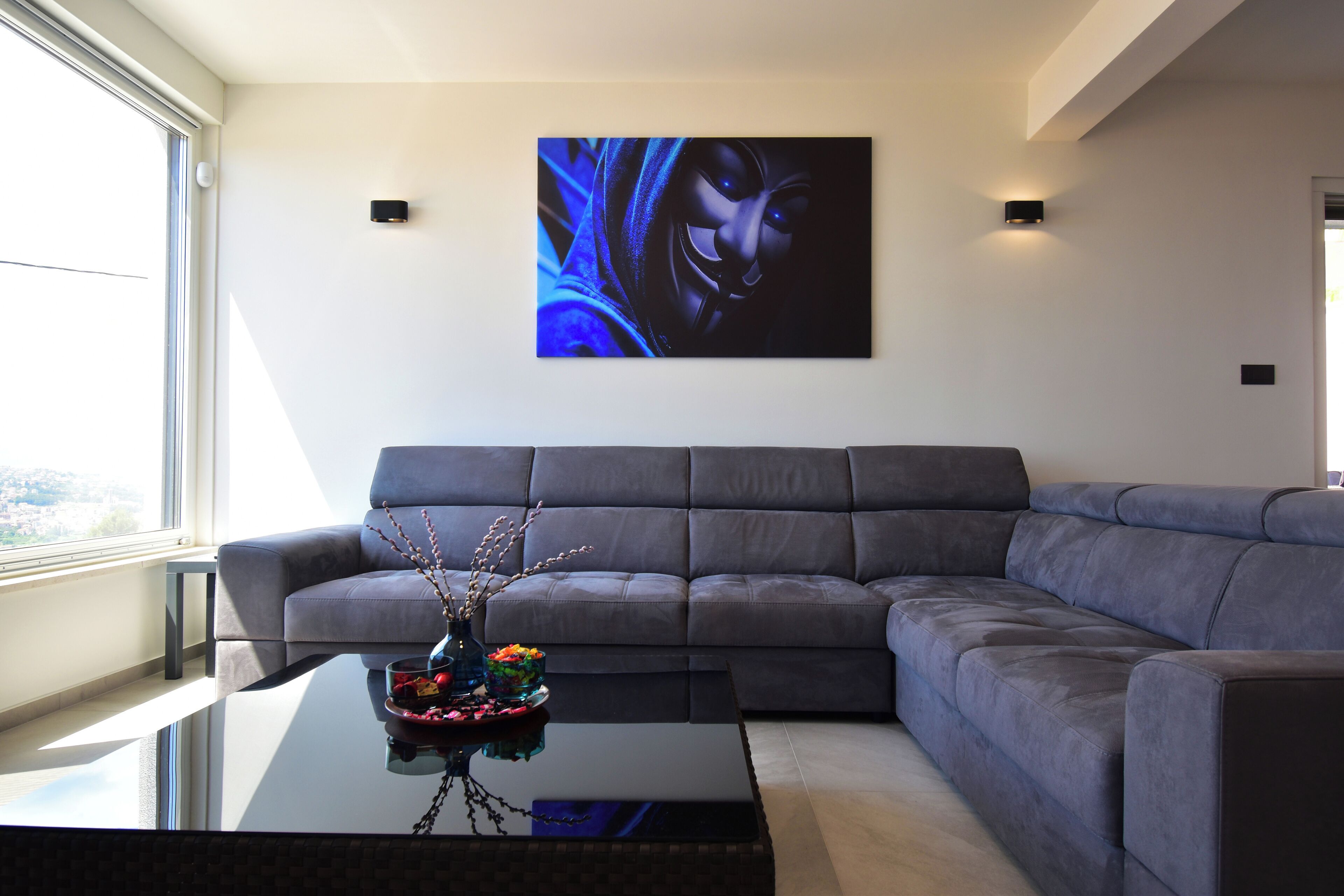 Una sala de estar moderna con un sofá seccional gris, una mesa de centro de vidrio con un florero y flores, y un cuadro abstracto grande en la pared.