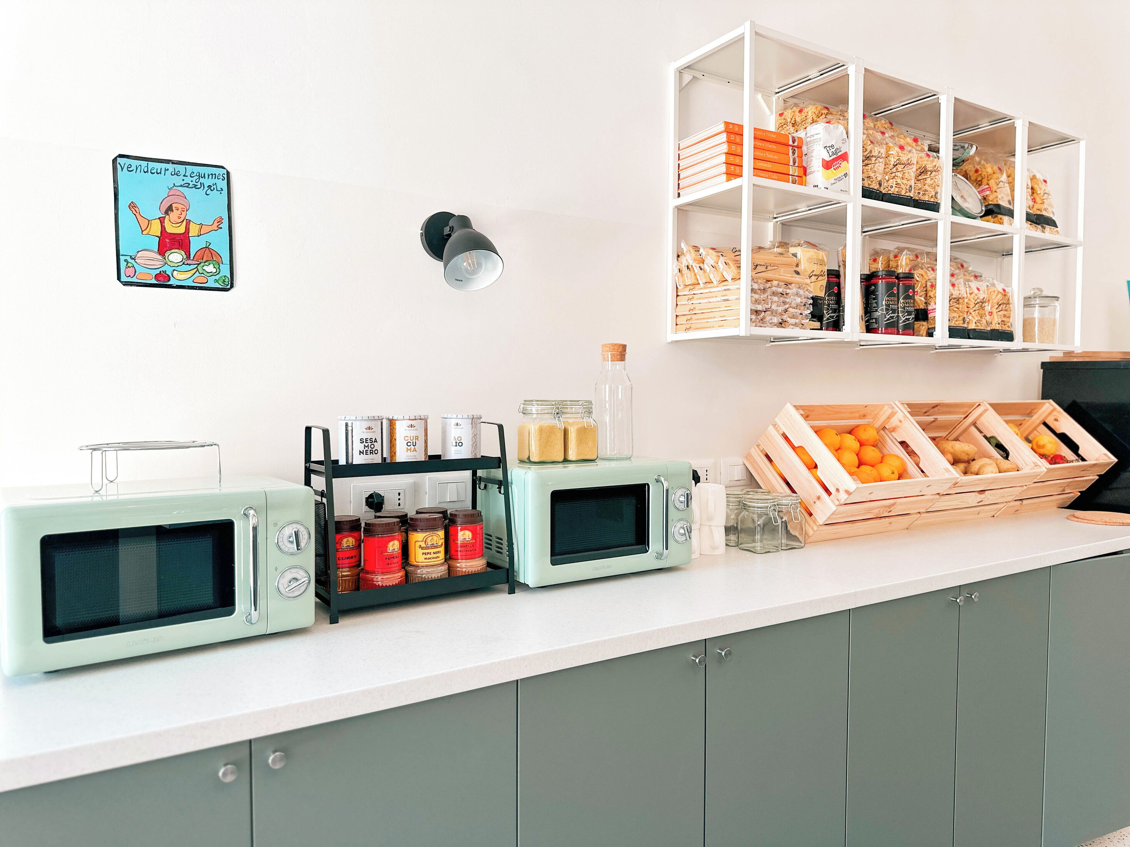 Un plan de travail de cuisine avec deux micro-ondes au style rétro, un étagère à épices et une caisse en bois remplie d’oranges.