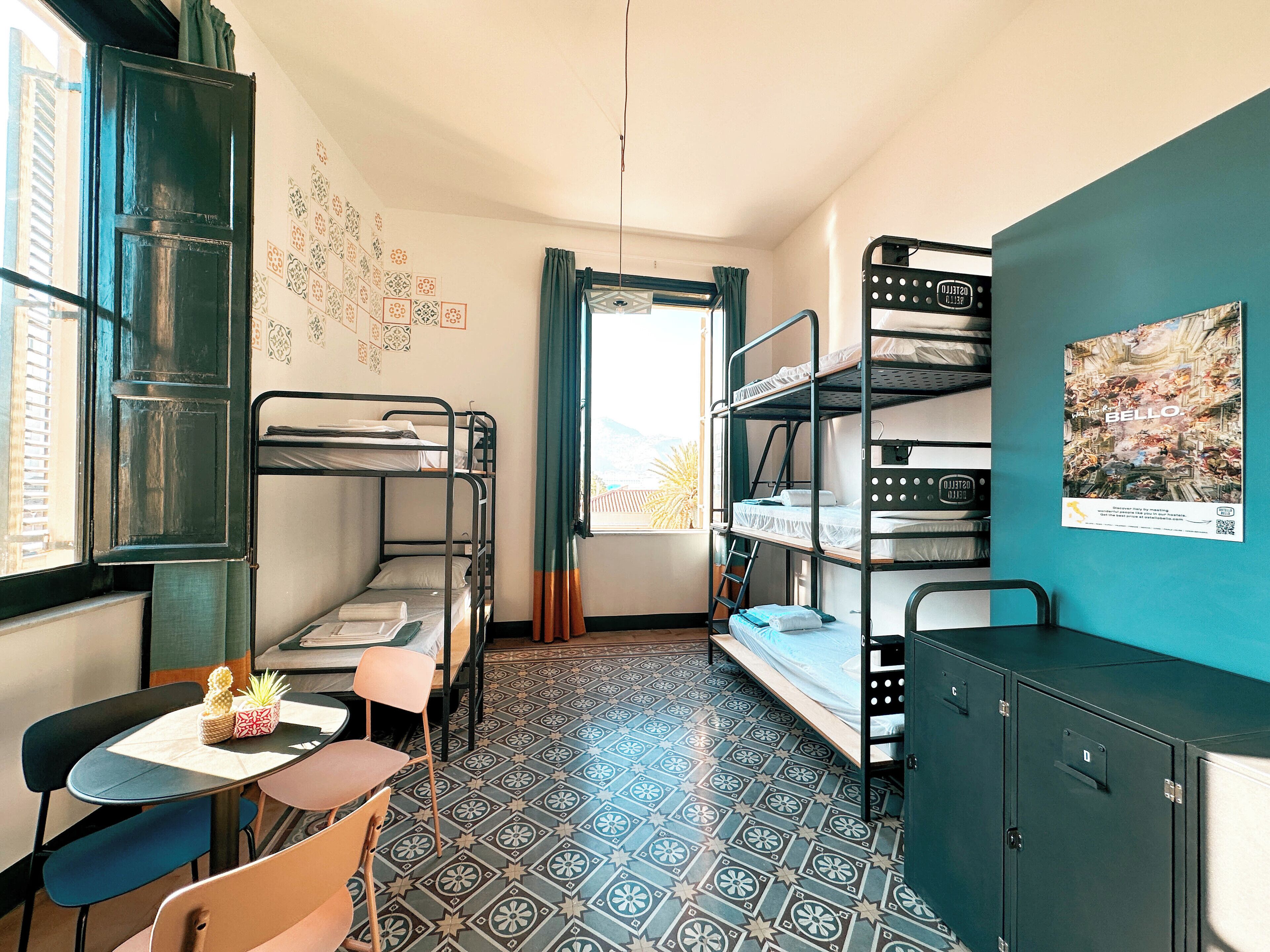 A dormitory room with bunk beds, a small table with chairs, a cabinet, and a window with curtains.