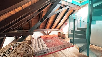 Een zolderkamer met houten balken, een rood geweven vloerkleed, een gewatteerde bank en een rieten stoel.