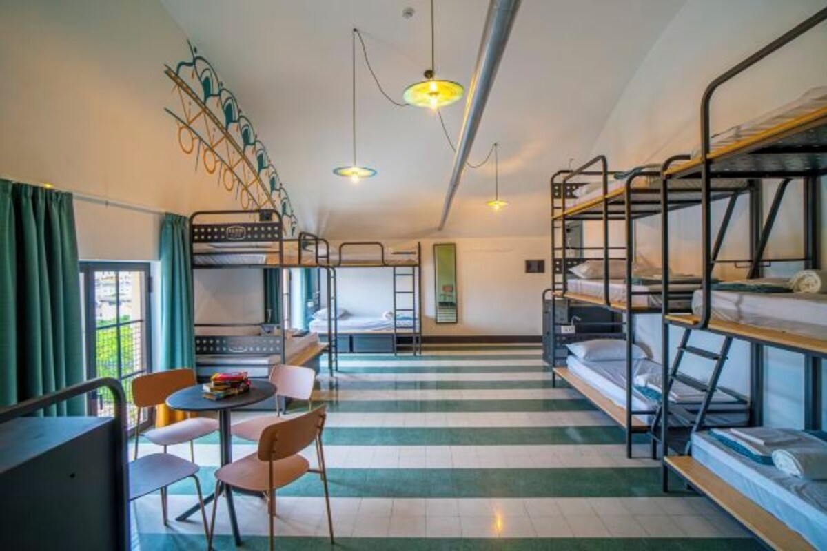 A dormitory room with bunk beds, a dining table with chairs, and a window with curtains.